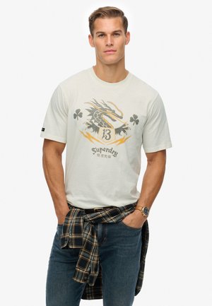 Cream short-sleeve t-shirt with a dragon graphic and "Superdry" text, paired with dark blue jeans and a black checkered flannel tied around the waist.
