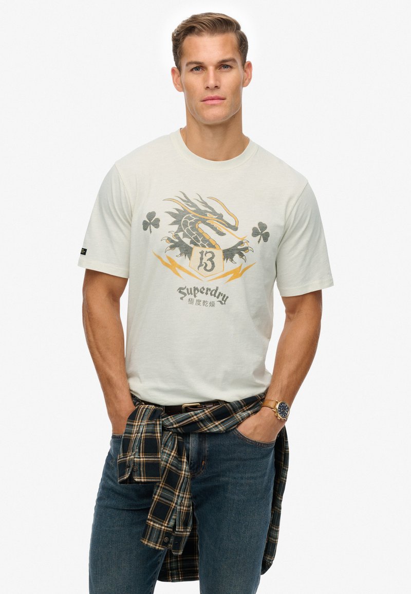 Crème kleurige t-shirt met korte mouwen, met een draak grafiek en "Superdry" tekst, gecombineerd met donkerblauwe jeans en een zwart-geruit flanelhemd dat om de taille is geknoopt.