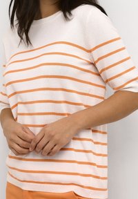 Vrouw in een witte korte mouw top met horizontale oranje strepen, staande tegen een witte muur, met de handen voorzichtig ineen gehaakt.