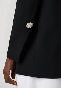 Jachetă neagră ajustată, cu un element decorativ texturat de argint pe mânecă, finisaj din țesătură netedă și siluetă ajustată.