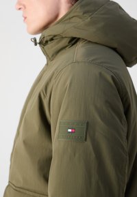 Olivengrøn hættejakke med en glat tekstur, der har et Tommy Hilfiger-logo på venstre ærme. Tætsiddende design med en diskret syning.