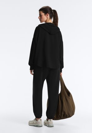 Žena s copom, oblečená v čiernej mikine s kapucňou a čiernych teplákoch, béžové tenisky, drží veľkú hnedú kabelku, stojí na jednoduchom pozadí.