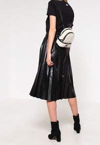 Svart plisserad kjol i glänsande läder, kombinerad med en svart figurnära topp och en liten, strukturerad krämfärgad crossbody-väska. Svarta ankelskor.