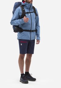 Hellblaues wasserdichtes Jackett mit attachierter Kapuze, marineblaue Shorts mit rotem Reißverschlussfach und dunklen Wanderschuhen mit Schnürsenkeln.
