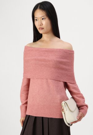 Pull rose décolleté avec texture côtelée, manches longues et taille ajustée ; associé à un sac à main crème.
