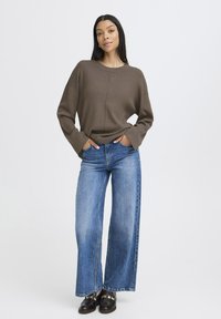 Pull marron côtelé avec des épaules tombantes, associé à un jean bleu clair à jambe large et des mocassins noirs avec des accents dorés.