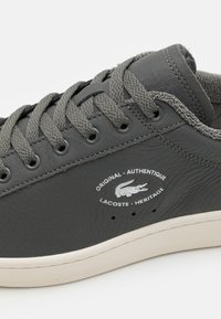Lacoste muške tenisice od tamnosive kože. Imaju zaobljeni dizajn s vezicama, teksturiranu površinu, gumu potplat i bijeli logotip s strane.