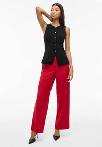 Svart ärmlös topp med V-formad nederkant och fem dekorativa knappar, kombinerad med vida röda byxor och svarta spetsiga klackar.