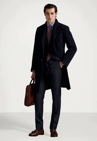 Άνδρας σε σκούρο ναυτικό παλτό, με ριγέ πουκάμισο, κόκκινη πουά γραβάτα, ναυτικά παντελόνια, καφέ δερμάτινα παπούτσια, κρατώντας καφέ δερμάτινη τσάντα, στέκεται μπροστά σε λευκό φόντο.