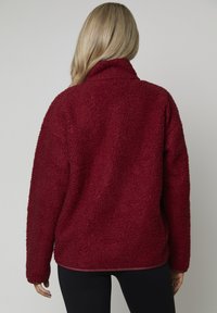 Burgundowa polarowa bluza z teksturowanym, miękkim wykończeniem. Posiada wysoki kołnierz i długie rękawy, zaprojektowana z myślą o luźnym kroju.
