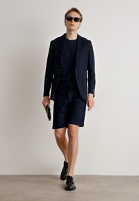 Blazer blu navy sopra una maglietta nera, abbinato a pantaloni corti blu navy. Gli accessori includono occhiali da sole e una pochette nera. Scarpe nere completano il look.