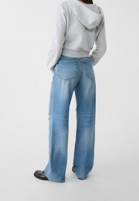 Vaaleanharmaa lyhyt huppari yhdistettynä leveälahkeisiin sinisiin farkkuihin. Farkuissa on hieman kulunut pinta ja näkyvää kulutusta lahkeen reunassa.