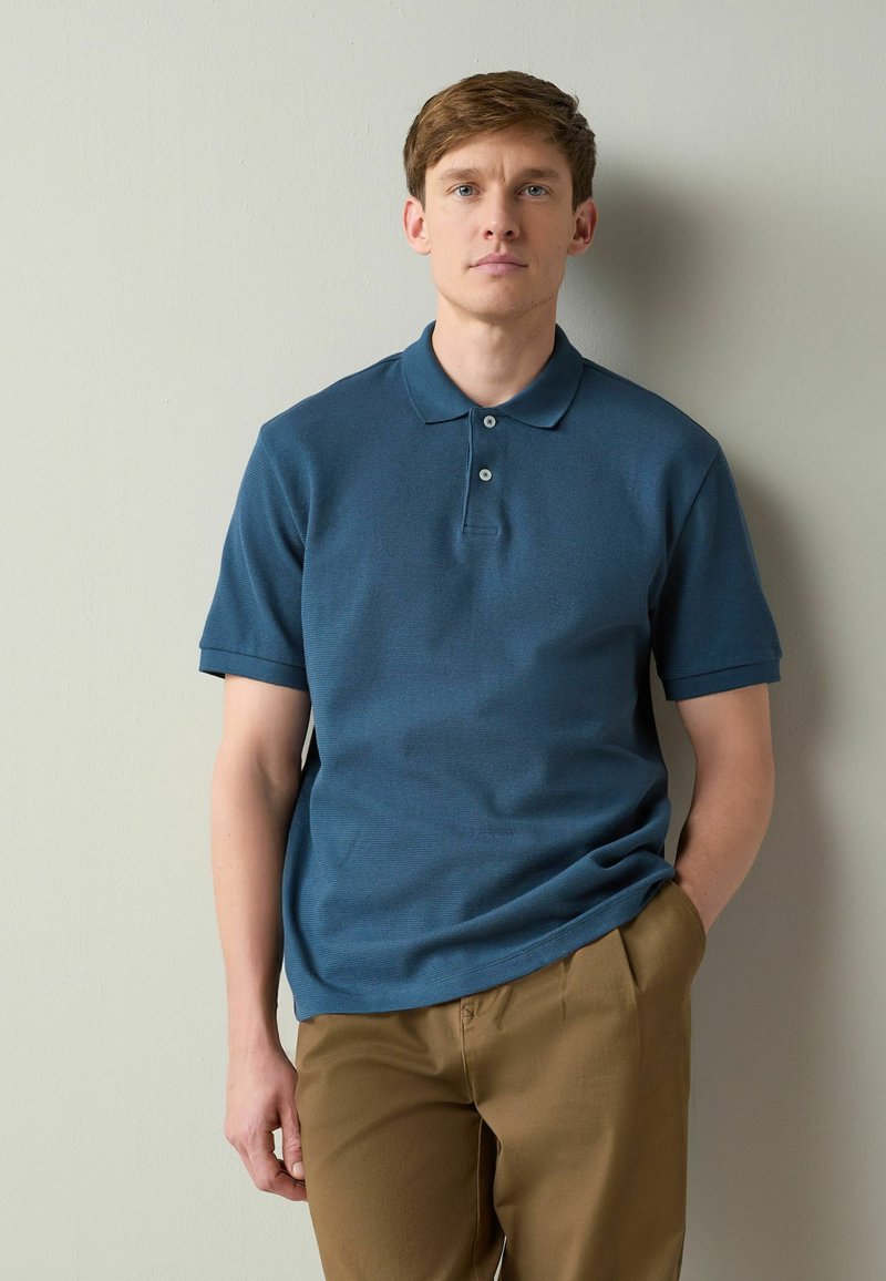 Tânăr cu păr scurt castaniu, purtând un tricou polo albastru și pantaloni maro, stând în picioare în fața unui perete simplu de culoare deschisă.