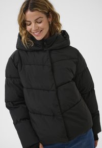 Schwarze Daunenjacke mit Kapuze, ausgestattet mit gesteppten Bereichen und einem eleganten Reißverschluss. Glattes, leichtes Material mit lockerer Passform.