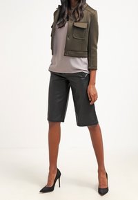 Olijfgroene cropped jack met twee voorkeurszakken, gecombineerd met een lichtgrijze top en zwarte nep leren bermuda shorts. Zwarte hoge hakken.