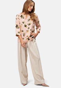 Donna con capelli lunghi che indossa una blusa floreale rosa con maniche a tre quarti, pantaloni beige a gamba larga e tacchi a punta, in piedi con una mano in tasca.