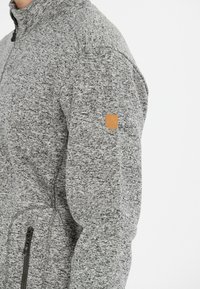 Grå fleecejakke med høj krave, der har tekstureret stof, en lynlåslomme på forsiden og et brunt læderlogo-patch på ærmet.