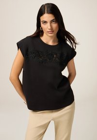 Maglietta nera in cotone con maniche corte, caratterizzata da un elegante design floreale in pizzo nero sul petto. Modello semplice e dalla vestibilità rilassata.