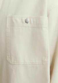 Cream-colored fabric with a textured finish. Features a chest pocket secured by a silver snap button. Clean stitching details visible.