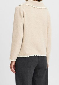 Pull en laine beige avec un col et un ourlet festonnés, doté de manches longues. Porté avec un pantalon gris foncé, présentant une finition douce et texturée.