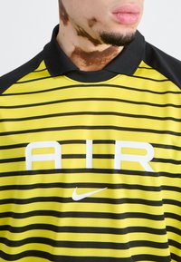 Dzeltena un melna svītrainā polo krekla ar melnu apkakli, kurā ir lieli baltie "AIR" burti un mazs balts Nike logotips. Gluda, sportiska auduma.