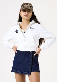 Sudadera blanca con cremallera y una estrella negra y logo, combinada con una falda de mezclilla oscura. La gorra presenta un patrón de estampado de leopardo.