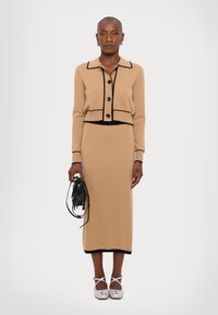 Beige knitted cardigan with black trim, button fastenings, paired with a matching long skirt. Silver flat shoes and a black bag with fringe detail.