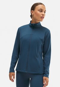 Chaqueta con cremallera en verde azulado oscuro, con mangas largas, cuello alto y bolsillos laterales. Fabricada en material liso y elástico con un diseño ajustado.