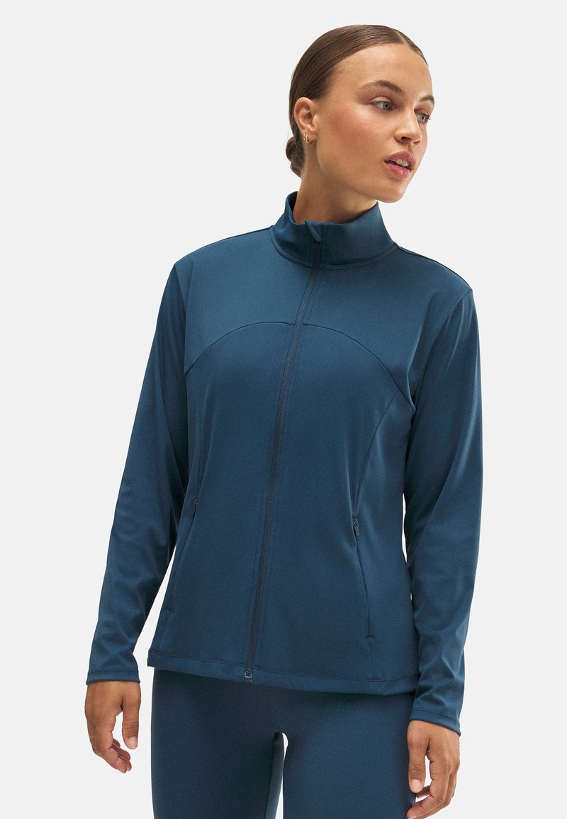 Chaqueta con cremallera en verde azulado oscuro, con mangas largas, cuello alto y bolsillos laterales. Fabricada en material liso y elástico con un diseño ajustado.