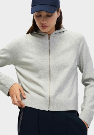 Felpa grigia con zip, con polsini e orlo a costine, dotata di cappuccio e texture liscia. Indossata sopra pantaloni scuri, il modello indossa un cappellino blu navy.