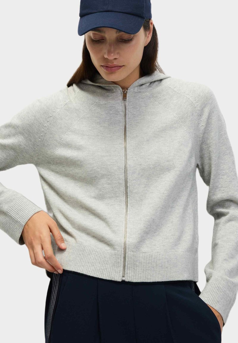 Grauer Reißverschluss-Hoodie mit gerippten Bündchen und Saum, ausgestattet mit Kapuze und glatter Textur. Über dunklen Hosen getragen, trägt das Modell eine marineblaue Mütze.