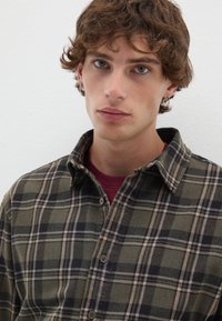 Camicia in flanella con un motivo a quadri verde e nero, colletto a bottoni e una texture morbida. Indossata sopra una maglia a girocollo bordeaux.