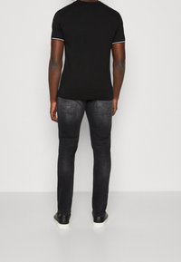 Camiseta negra de manga corta con ribete blanco, combinada con jeans oscuros de corte ajustado y zapatillas negras, vista desde la parte de atrás.