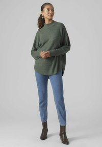 Dark green knitted sweater with a high neckline, loose fit, and ribbed cuffs, paired with light blue straight-leg jeans and brown ankle boots.