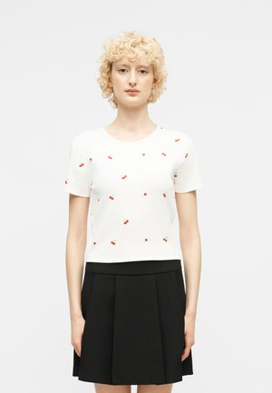 Ξανθιά γυναίκα με σγουρά μαλλιά φοράει λευκό τοπ με κοντά μανίκια και μικρά τυπώματα κερασιών, καθώς και μαύρη πλισέ φούστα, στέκεται μπροστά σε απλό φόντο.