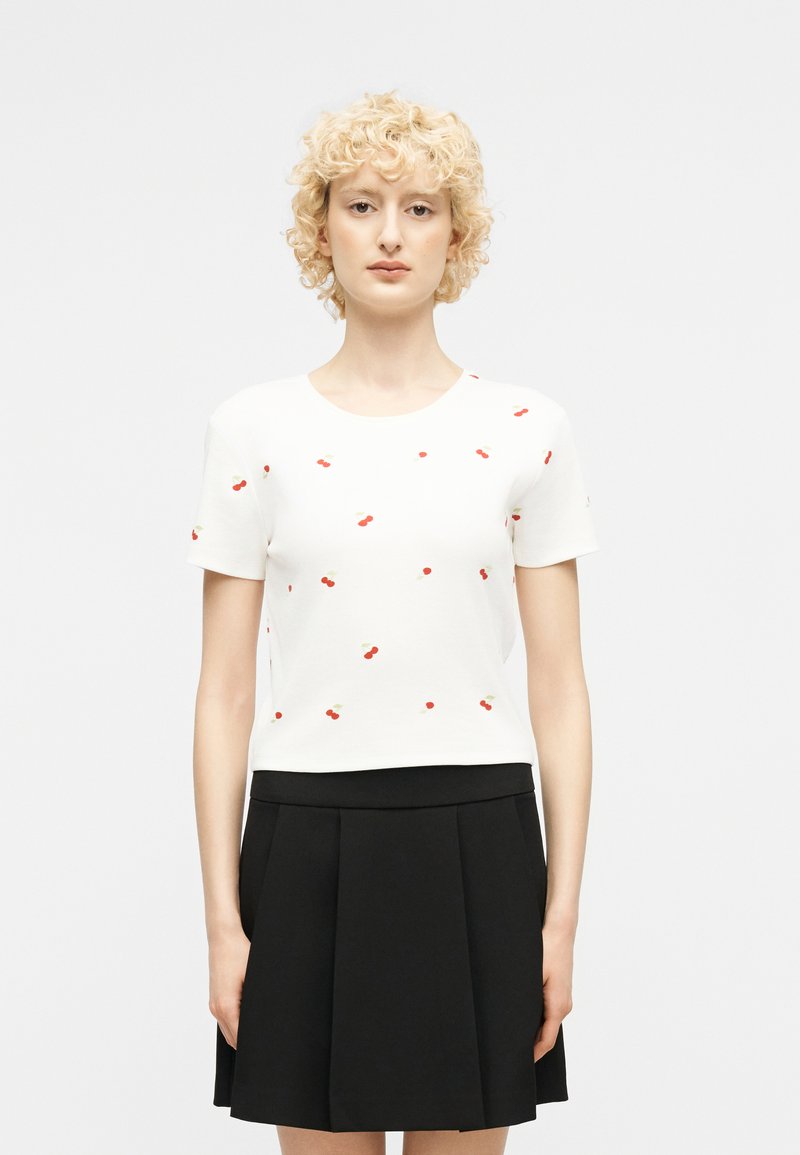 Ξανθιά γυναίκα με σγουρά μαλλιά φοράει λευκό τοπ με κοντά μανίκια και μικρά τυπώματα κερασιών, καθώς και μαύρη πλισέ φούστα, στέκεται μπροστά σε απλό φόντο.