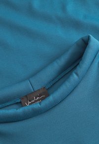 Tricou teal cu o textură moale și netedă; are un guler rotund și un etichetă cu "Vaschi" imprimată cu font alb cursiv.