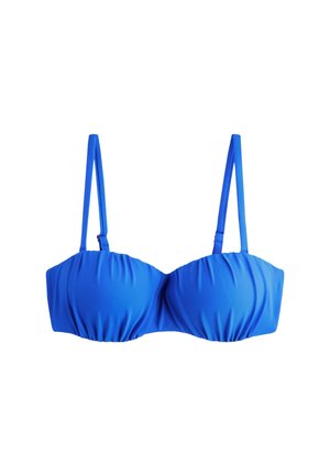 Donna con lunghi capelli mossi che indossa un bikini blu, in piedi accanto a un muro bianco con texture, tiene un asciugamano e guarda in basso verso sinistra.