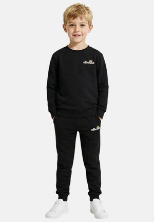 Jeune garçon debout, portant un sweat-shirt et un pantalon de survêtement noirs de la marque ellesse, avec des baskets blanches, mains dans les poches, souriant à la caméra.