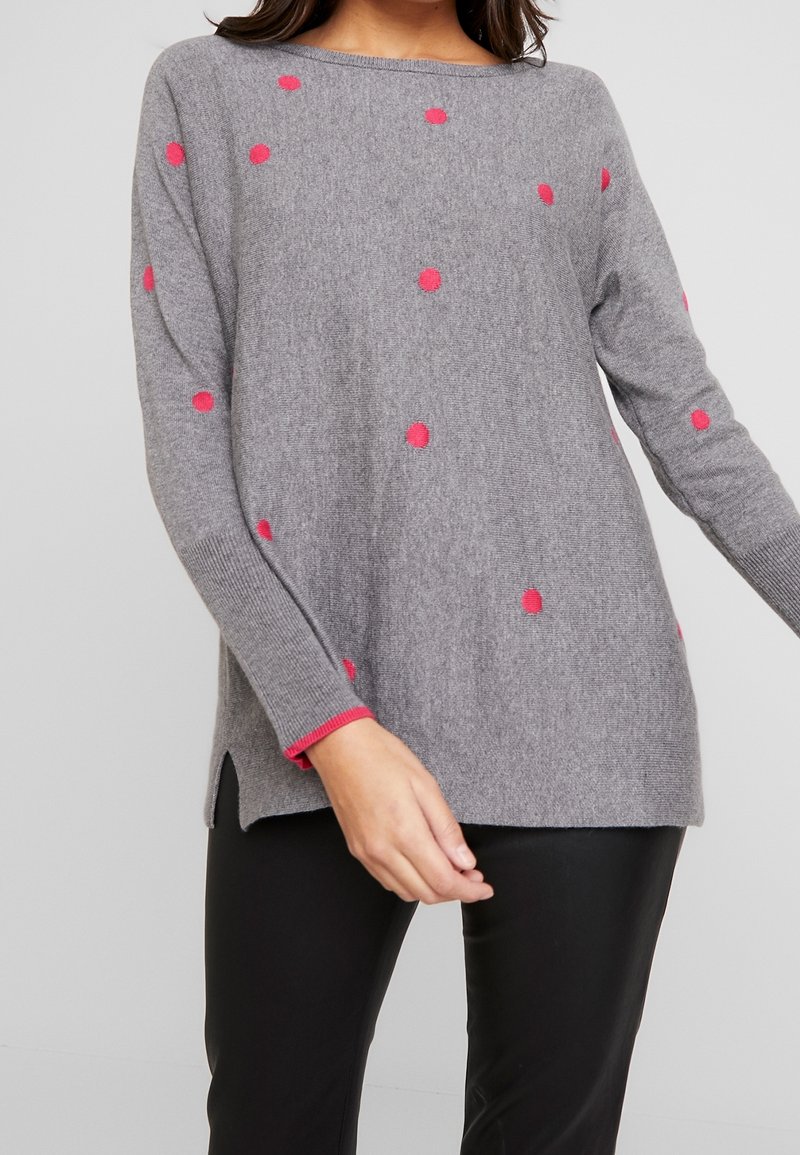 Femme portant un pull gris ample avec de petits pois roses et un pantalon noir, debout devant un fond clair uni.