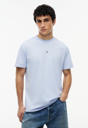 Junger Mann mit lockigem dunklem Haar, der ein hellblaues "Tommy Jeans"-T-Shirt und blaue Jeans trägt, stehend mit einer Hand in der Hosentasche.