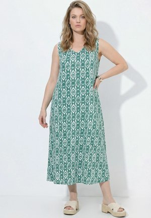 Kvinde med bølget blondt hår iført en ærmeløs midi-kjole med grønne og hvide mønstre og beige kilehælssandaler, stående med den ene hånd på hoften.