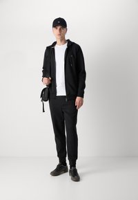 Felpa nera con zip sopra una maglietta bianca, abbinata a pantaloni sportivi neri e sneakers. Un cappellino nero e una piccola borsa a tracolla completano l'outfit.