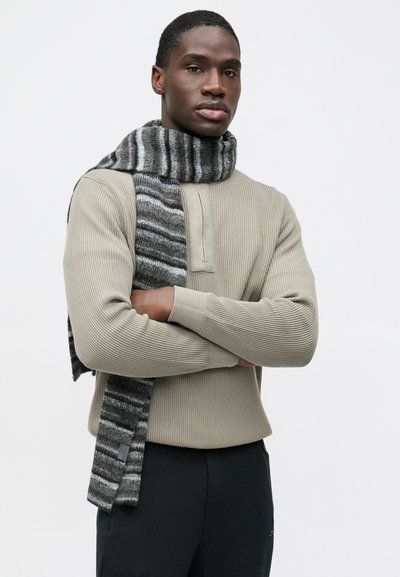 Γκρι ριγέ κασκόλ με απαλό ύφασμα που είναι φορεμένο γύρω από το λαιμό ενός ατόμου, σε συνδυασμό με ένα ανοιχτό μπεζ ριμπ πλεκτό πουλόβερ.