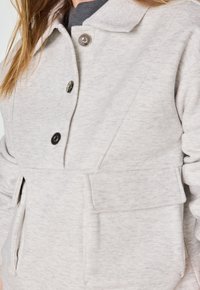 Sweatshirt gris clair, texturé, avec un col, deux poches avant et fermeture à boutons-pression. Le matériau semble doux et décontracté.