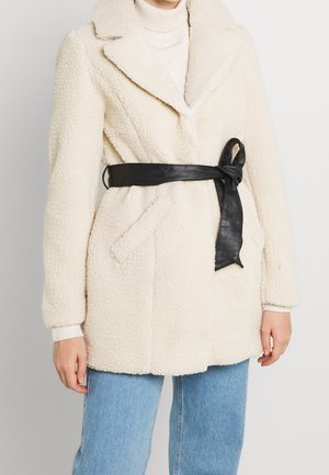 Femme portant un manteau sherpa crème avec ceinture en cuir noire, un col roulé blanc et un jean bleu, debout devant un fond clair uni.