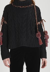 Maglione nero a maglia con colletto alto, caratterizzato da motivi a treccia, dettagli intrecciati e decorazioni a pompon sulle maniche.