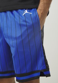 Kék kosárlabda sort fekete függőleges csíkokkal, hálós anyagból, fekete oldalsó kiegészítőkkel és fehér Jumpman logóval a derékpánt közelében.