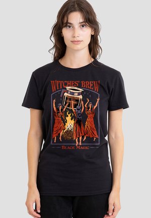 Woman wearing a black T-shirt with an illustration of three witches around a fire lifting a large coffee pot labeled "coffee."