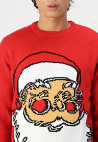 Pull en laine rouge avec un grand motif de Noël représentant le Père Noël. Le design comprend des accents noirs et des détails blancs, mettant en avant le thème festif.